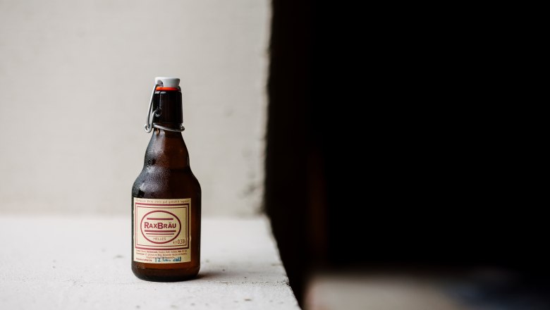 A bottle of RaxBräu Helles Bier on a white surface against a dark background.