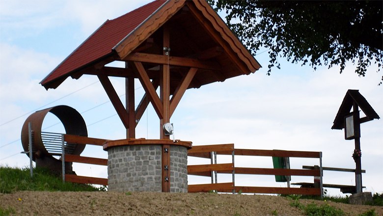 A covered fountain made of stone and wood with a fence next to it and a large metal ring in the background.