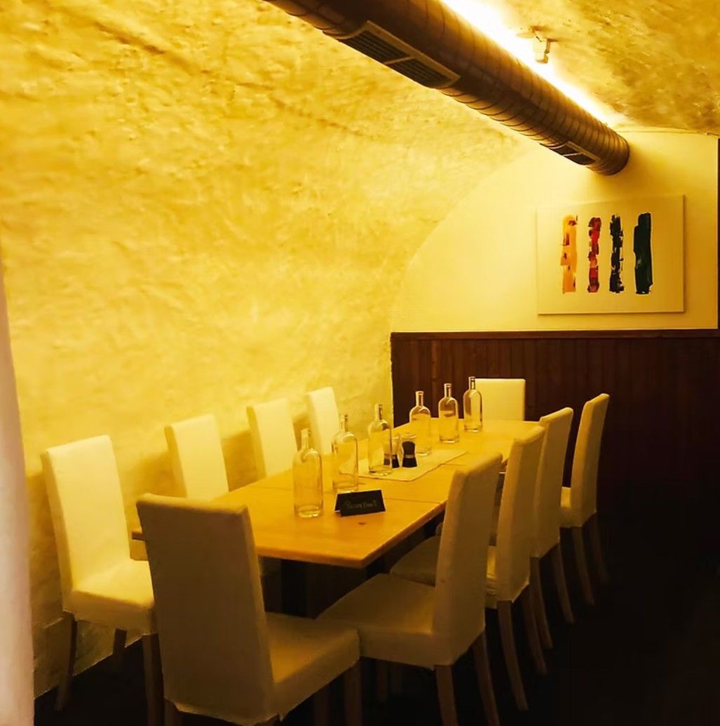 A long table with white chairs in a vaulted cellar, decorated with empty bottles and a modern painting on the wall.
