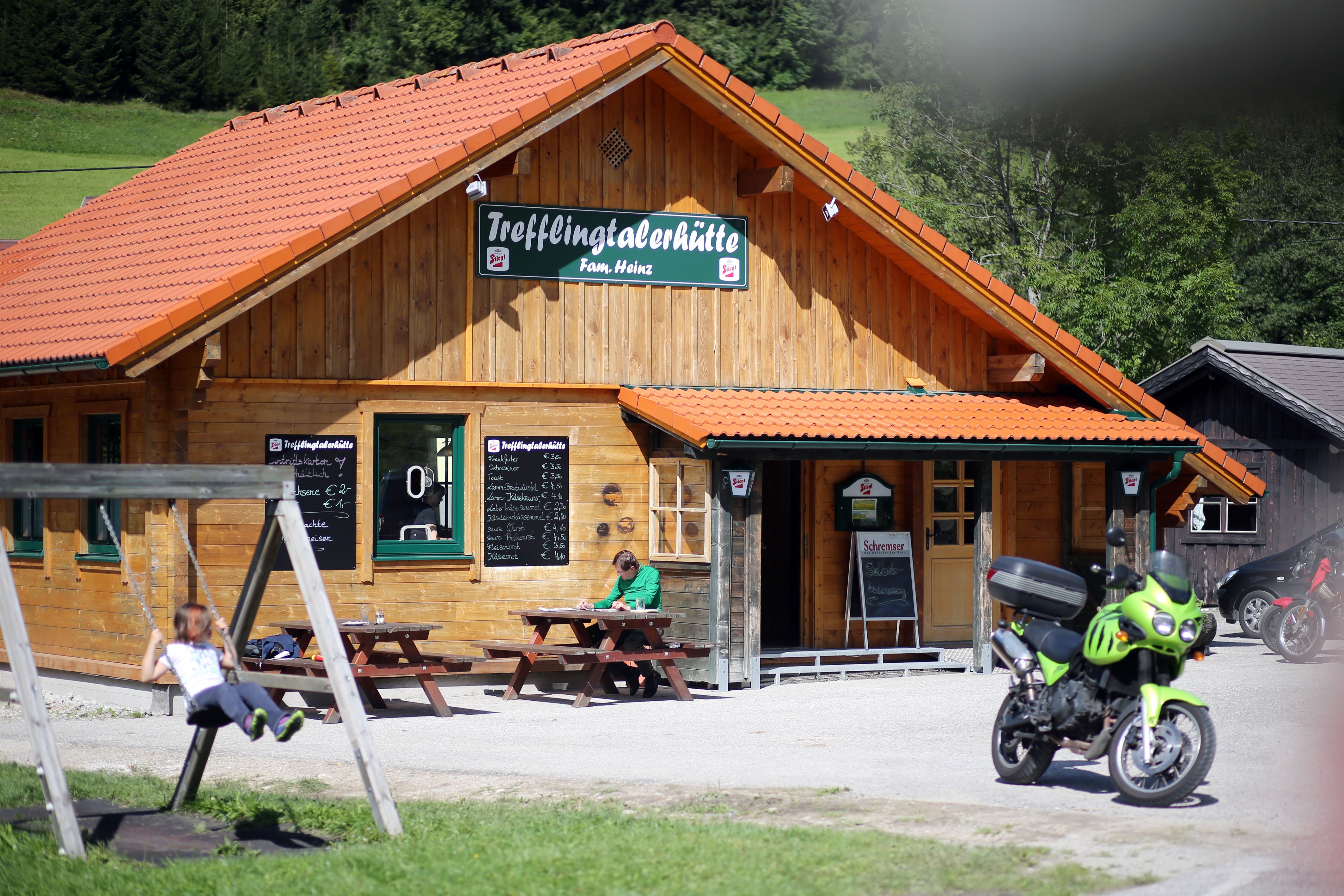 A wooden hut with the sign 'Trefflingtalerhütte', in front of it a swing, a motorcycle and a person at a table.