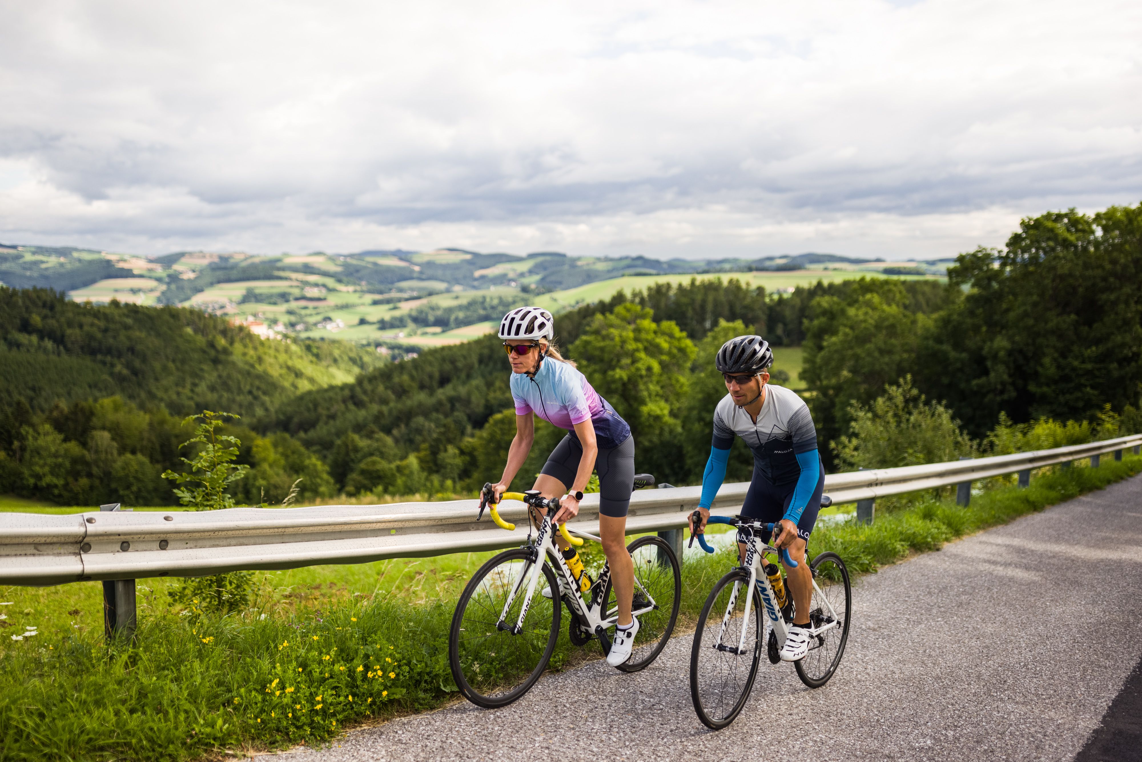 Two cyclists are riding on a road in a hilly landscape.