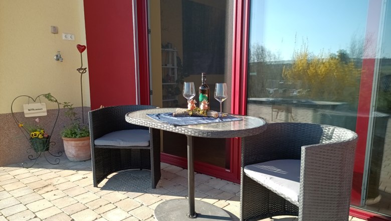 Terrace with table, two chairs, bottle of wine and glasses in front of a red door.