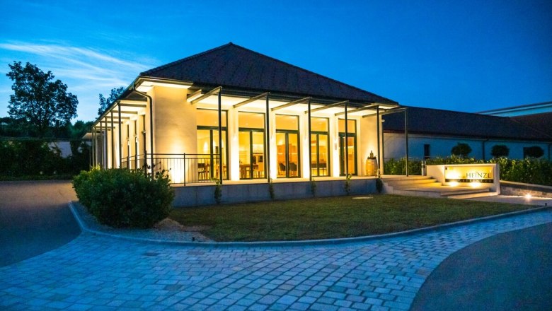A modern building with glass fronts, illuminated at dusk, surrounded by a paved path and green spaces.
