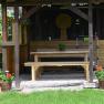 A rustic gazebo with a wooden table and benches, surrounded by plants and flowers in baskets.