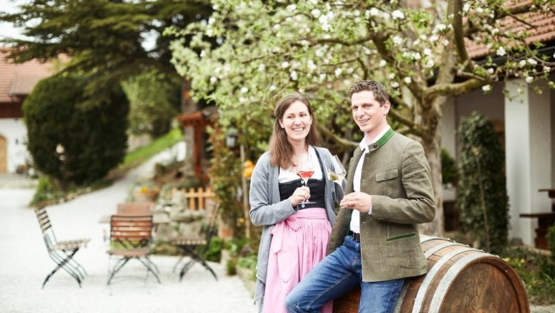 A couple in traditional dress stand smiling with wine glasses in front of a wine barrel outside.