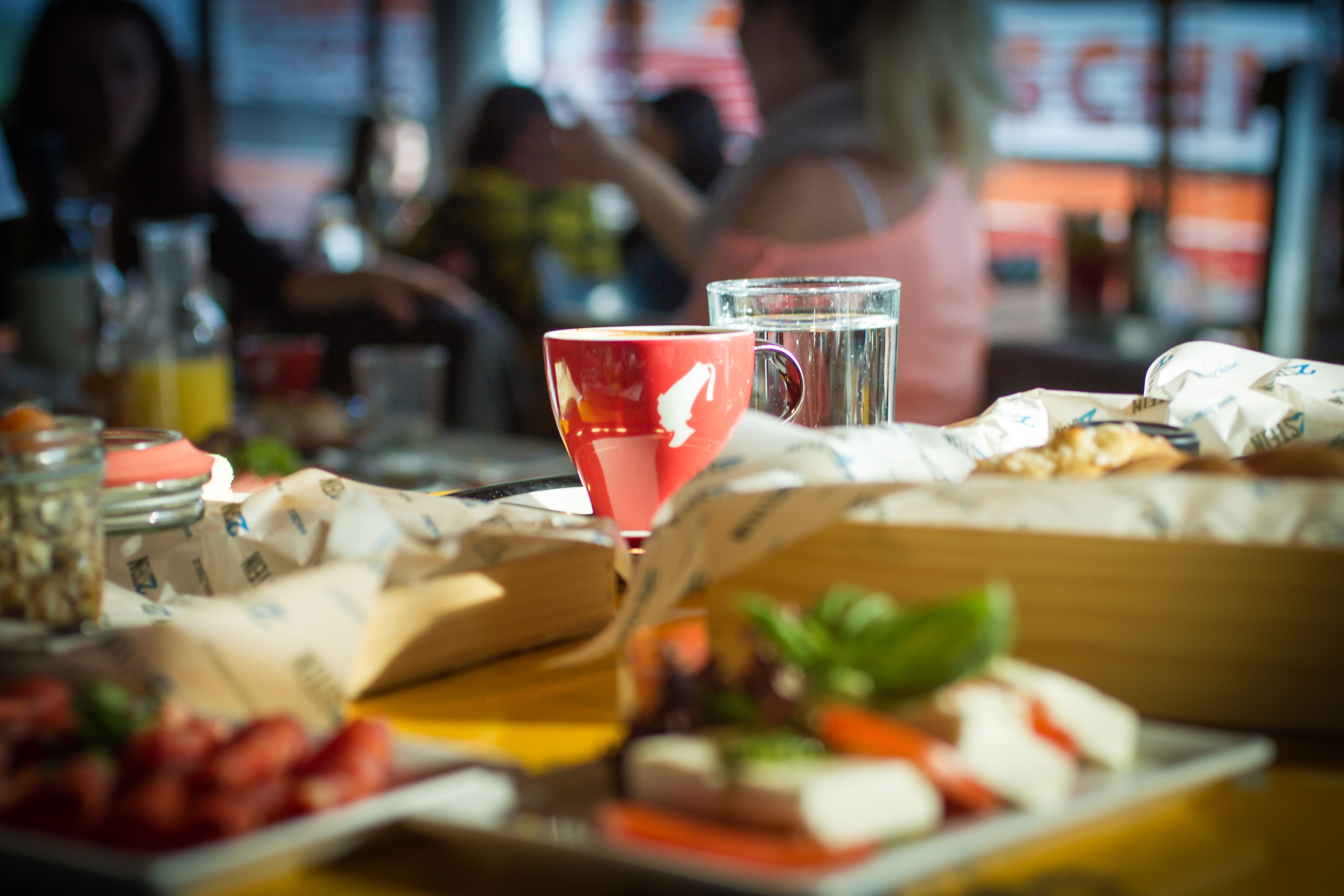 A red coffee mug and a glass of water on a table in a café, surrounded by food and drinks.