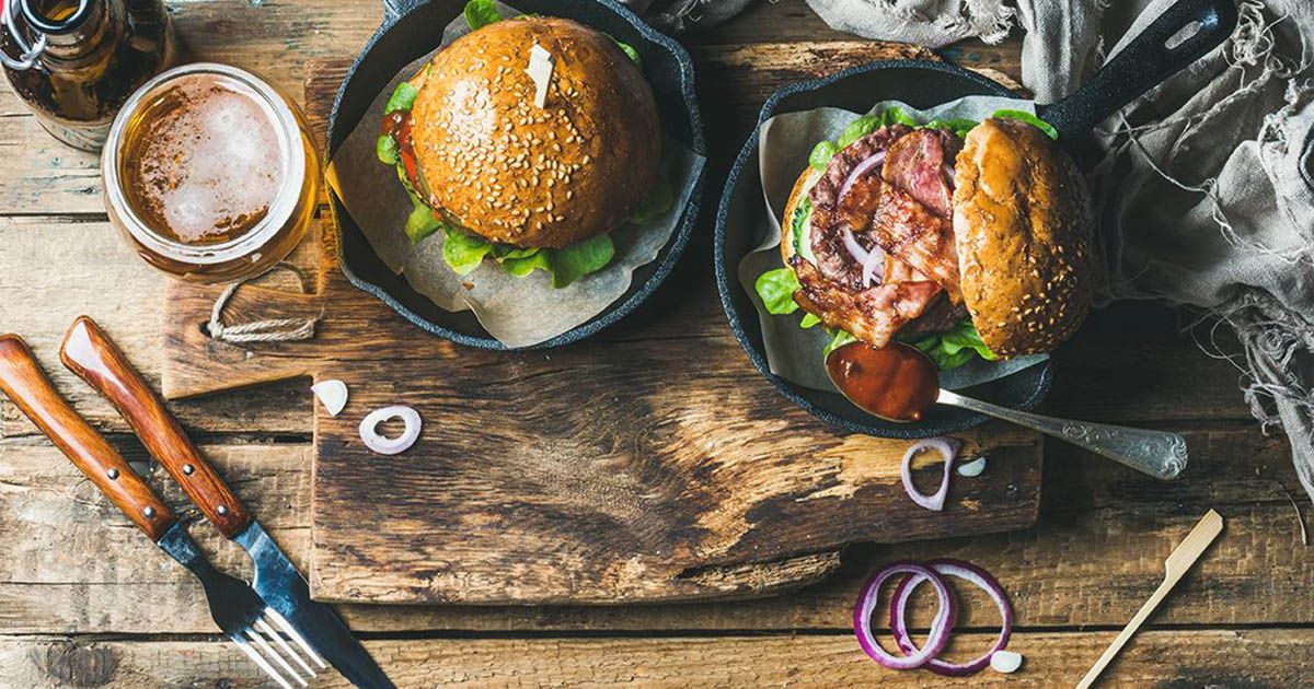 Two burgers in pans with beer and cutlery on a wooden table.