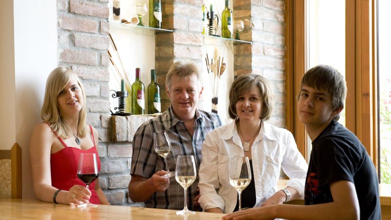Four people sit at a table with wine glasses, wine bottles on shelves in the background.