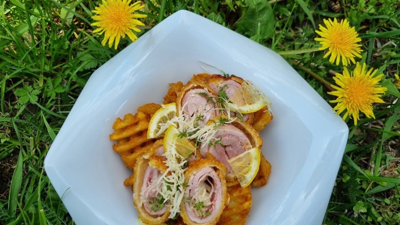 A plate of cordon rolls and waffle fries, garnished with lemon slices and cress, lies on a meadow of dandelions.