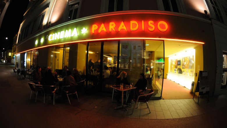 Exterior view of a movie theater called Cinema Paradiso at night with illuminated entrance and people at tables.
