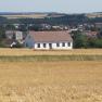 A white house with a red roof stands in a field, with a town and woods in the background.