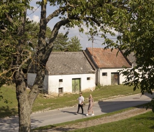 Two people walk past old buildings in a rural setting.