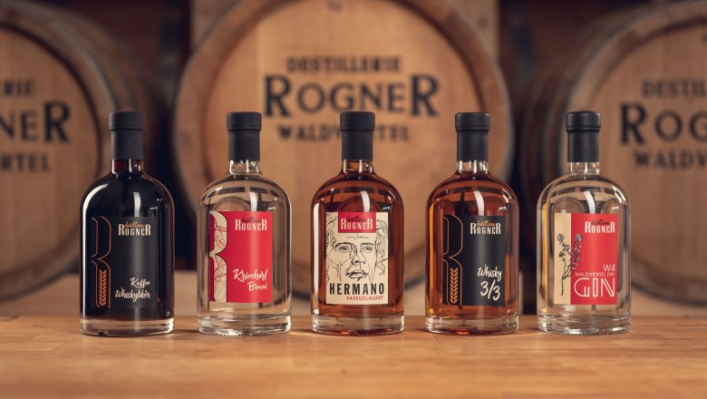 Five bottles of spirits from the Rogner distillery in front of wooden barrels.