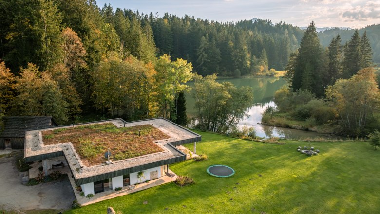Vacation home by the lake, © Niederösterreich Werbung / Maximilian Pawlikowsky Aerial view of a modern house with a green roof, surrounded by forest and a lake.