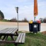 Picnic table and garbage can in a parking lot with trees in the background.