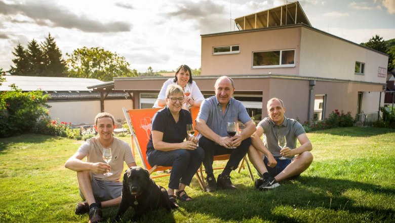 A family sits with a dog on a lawn in front of a modern house, holding glasses of wine.