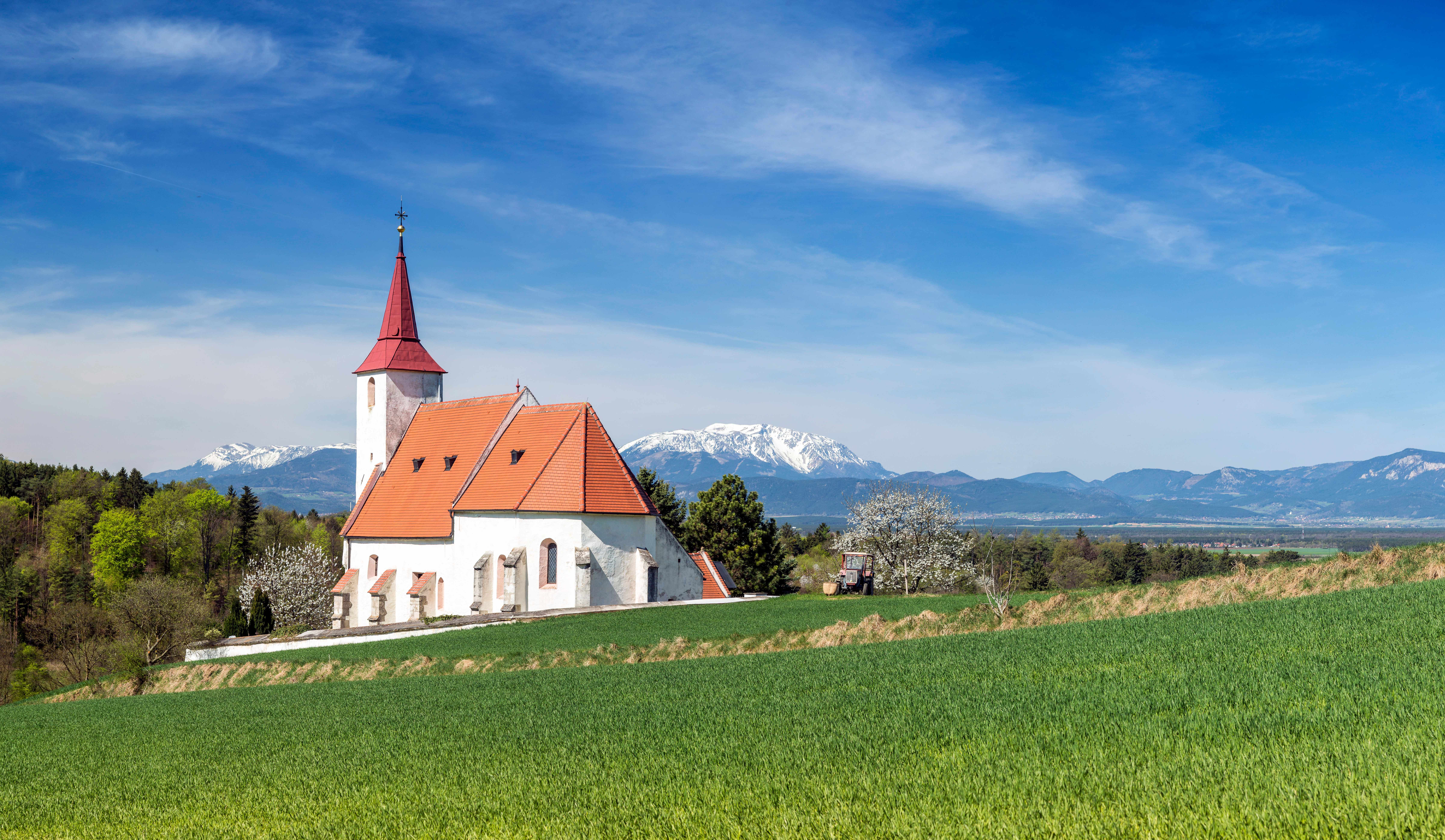 White Ofenbach parish church with red roof and church tower against a mountain backdrop with a green field in the foreground.