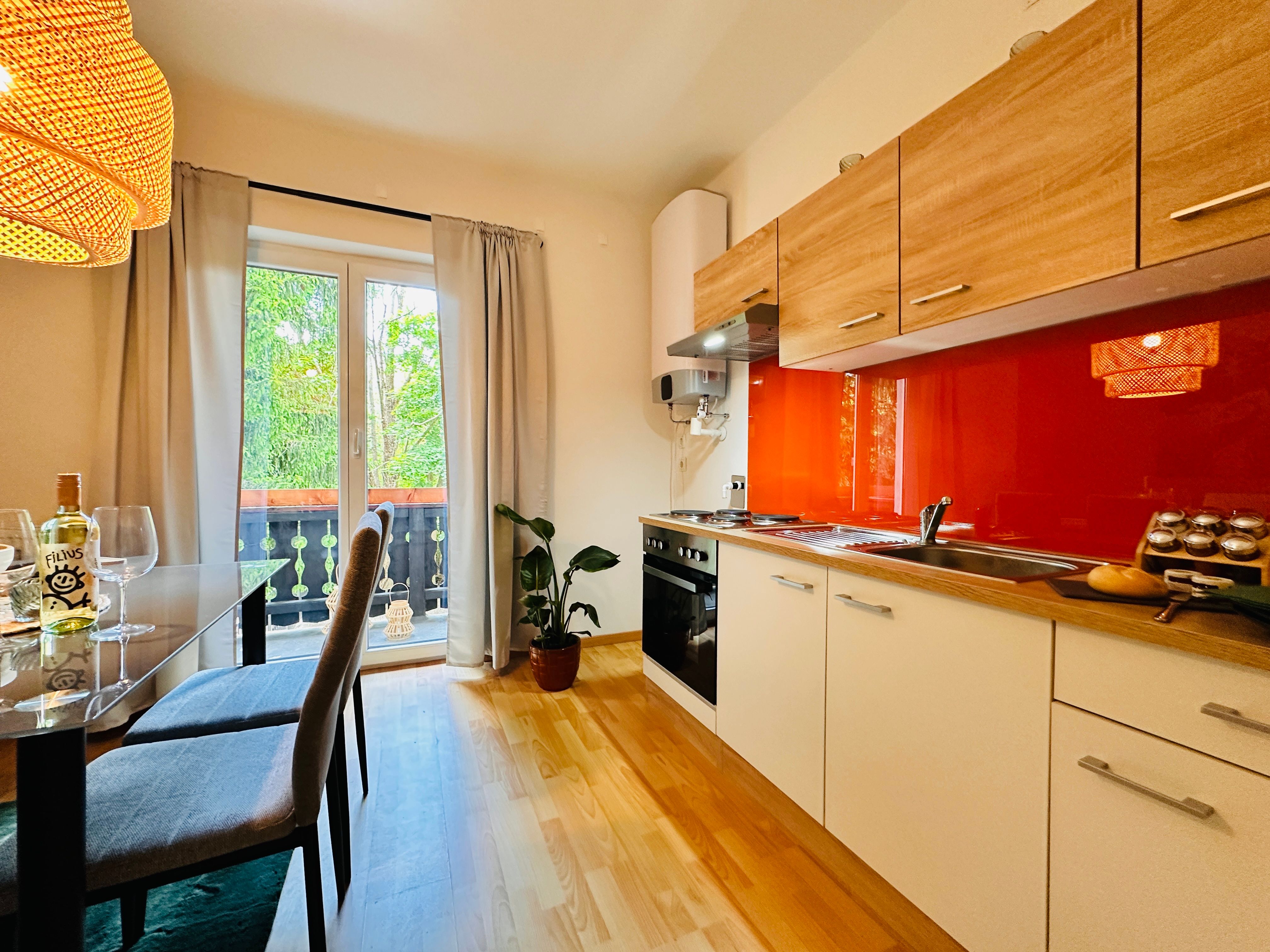Cozy kitchen with wooden furniture, red splashback and dining table. View of balcony with plants.