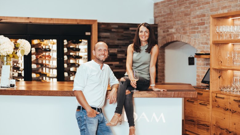Two people in a modern wine bar, a woman sitting on the counter, a man leaning against it. Wine racks and glasses in the background.