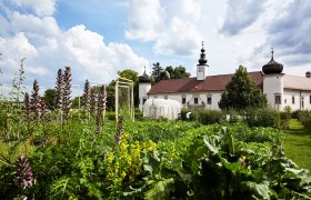 A monastery building with onion domes behind a lush garden under a blue sky.
