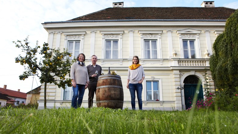 Three people stand in front of a historic building with a wine barrel in the foreground.