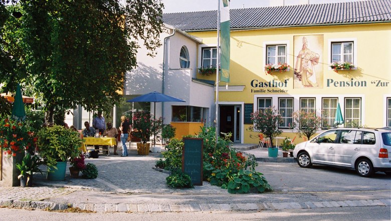 A yellow building with the inscription 'Gasthof-Pension Zur Hammerschmiede'. There are tables, chairs and a car in front of the building.