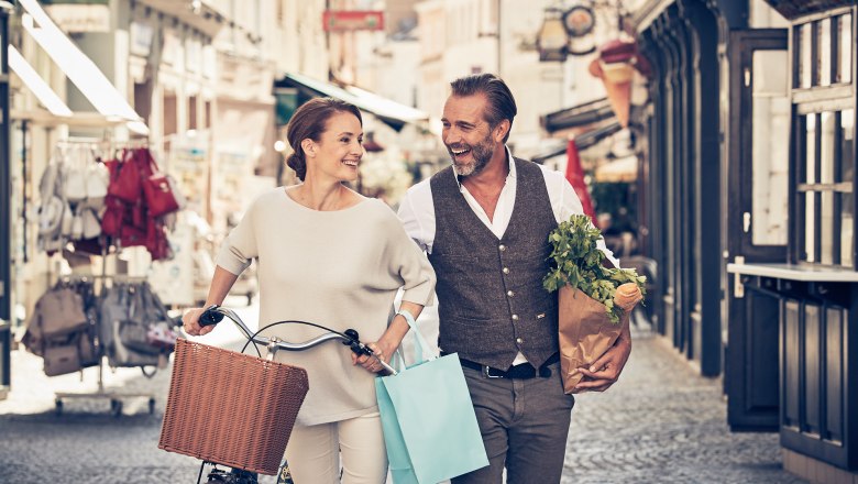 A smiling couple strolls through an old town with shopping bags and a bicycle.