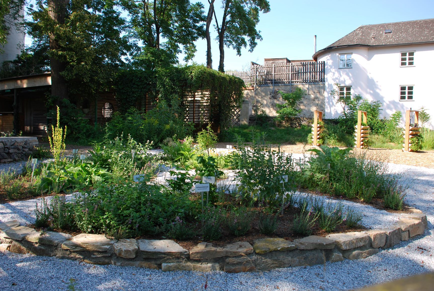A well-tended garden with various plants and signs, surrounded by trees and a building in the background.