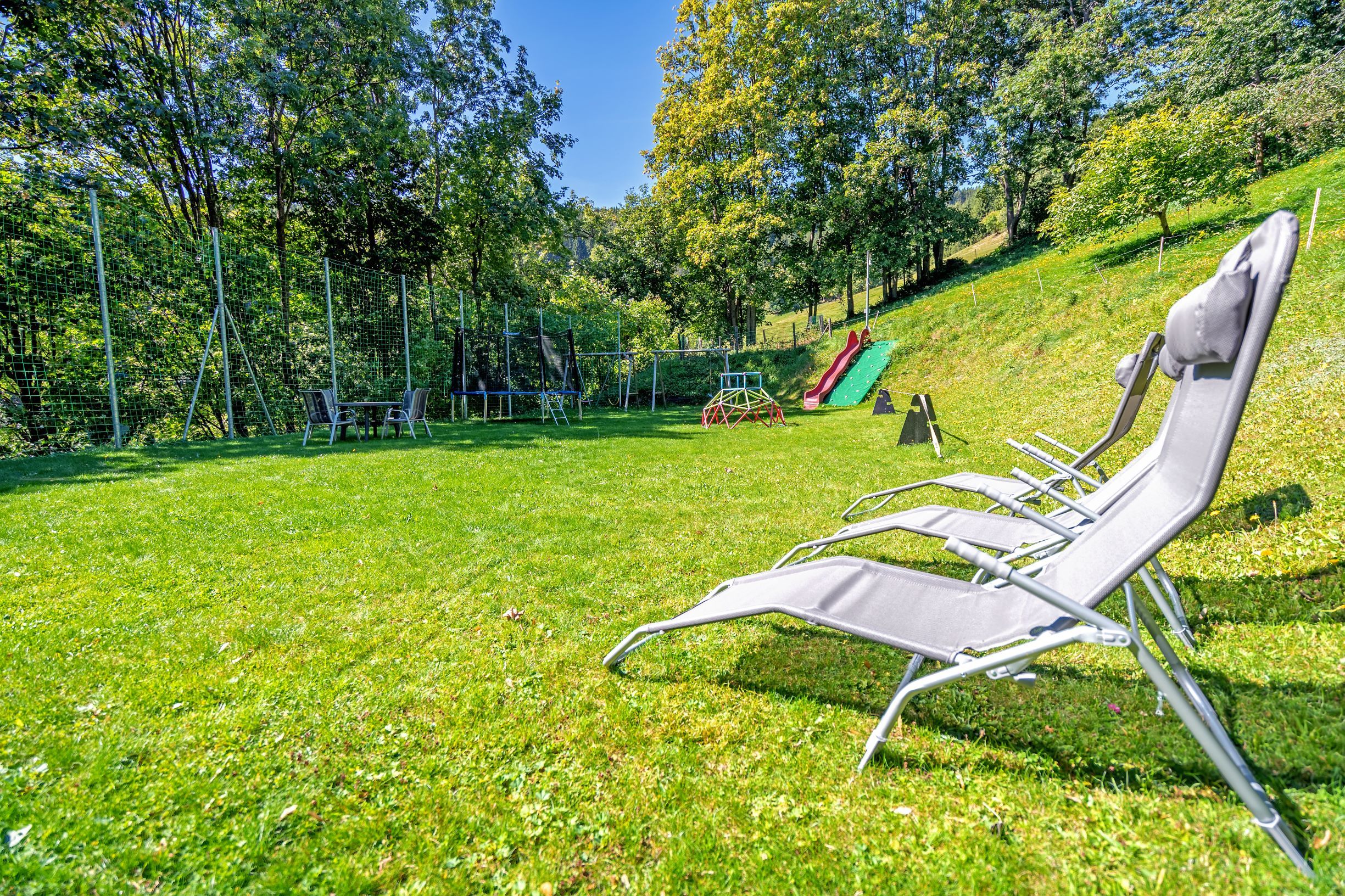 Playground with slide, trampoline and sun loungers on a meadow surrounded by trees.