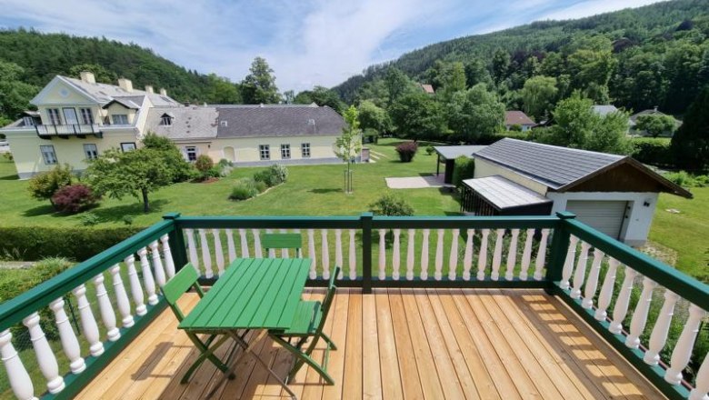 View from a balcony onto a garden with a green meadow and trees, with a large house and hills in the background.