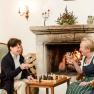 Two women are sitting in armchairs in front of a fireplace, drinking wine and playing chess.