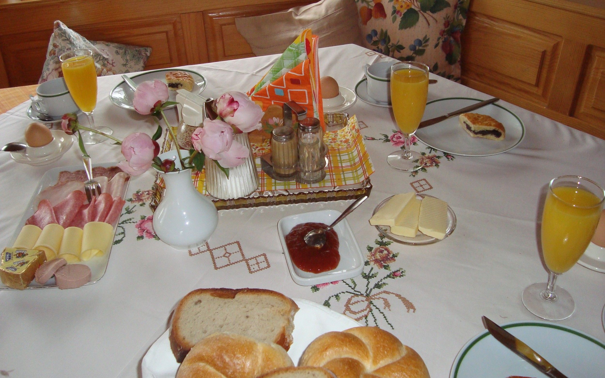 A laid breakfast table with bread rolls, cold cuts, cheese, jam, orange juice and flowers.