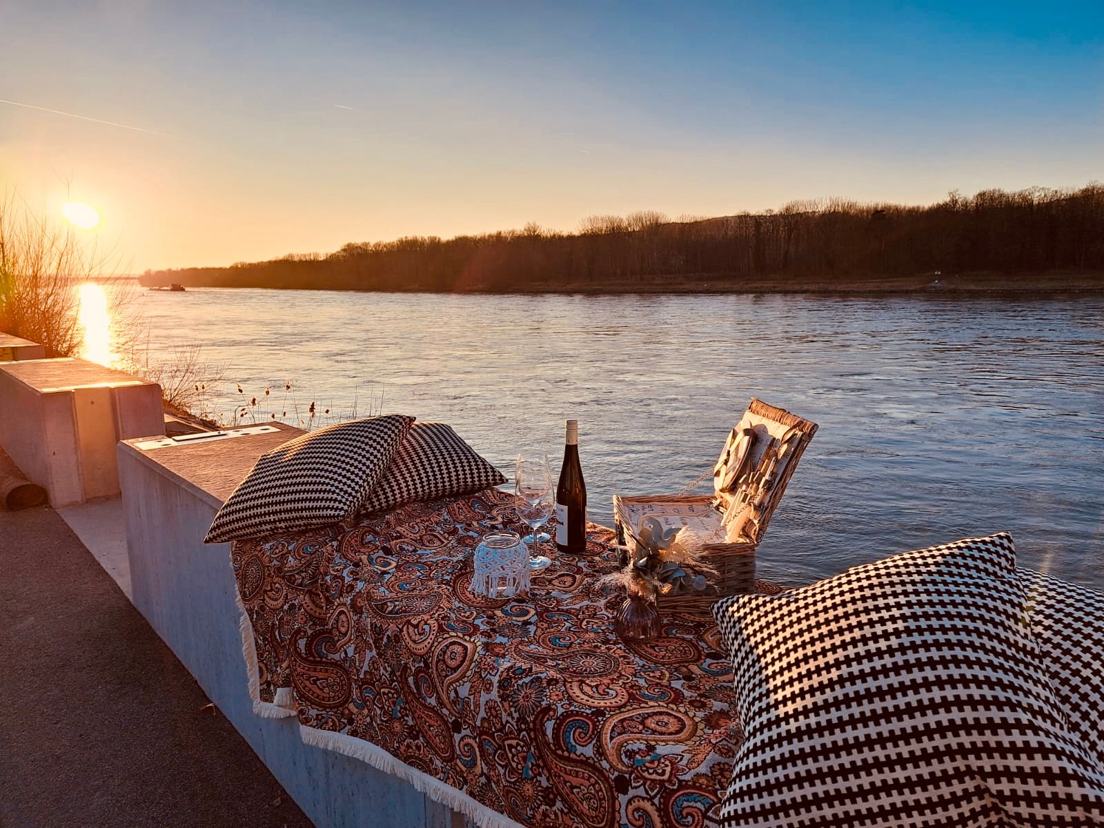 Picnic on the riverbank at sunset with a blanket, cushion, wine and picnic basket.