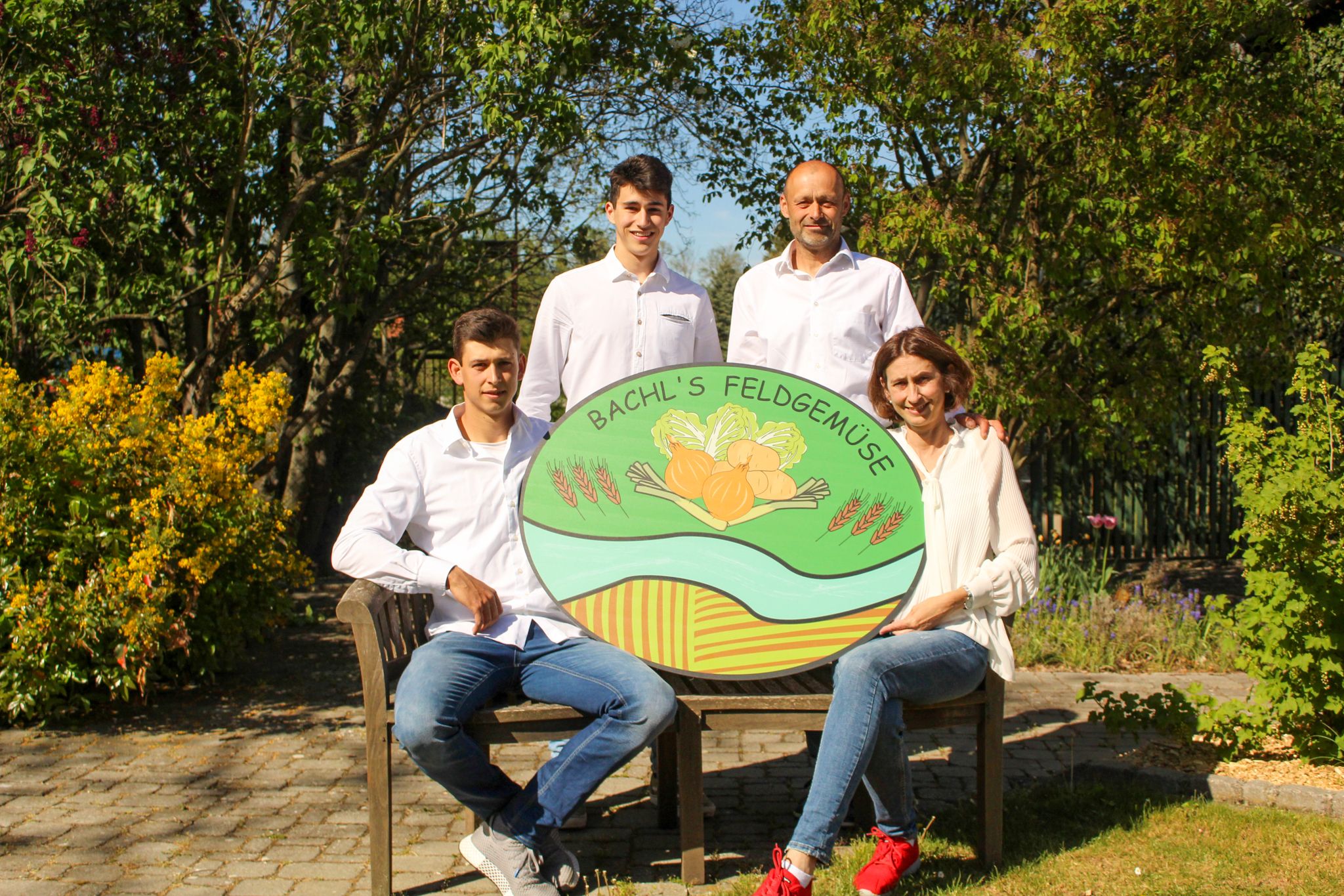 Four people pose with a sign for 'Bachl's Feldgemüse' in a garden.