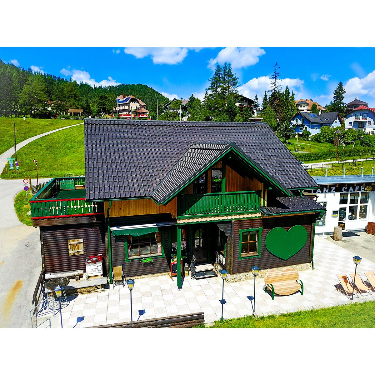 A traditional wooden house with a green heart motif and terrace in summer, surrounded by green hills and blue skies.