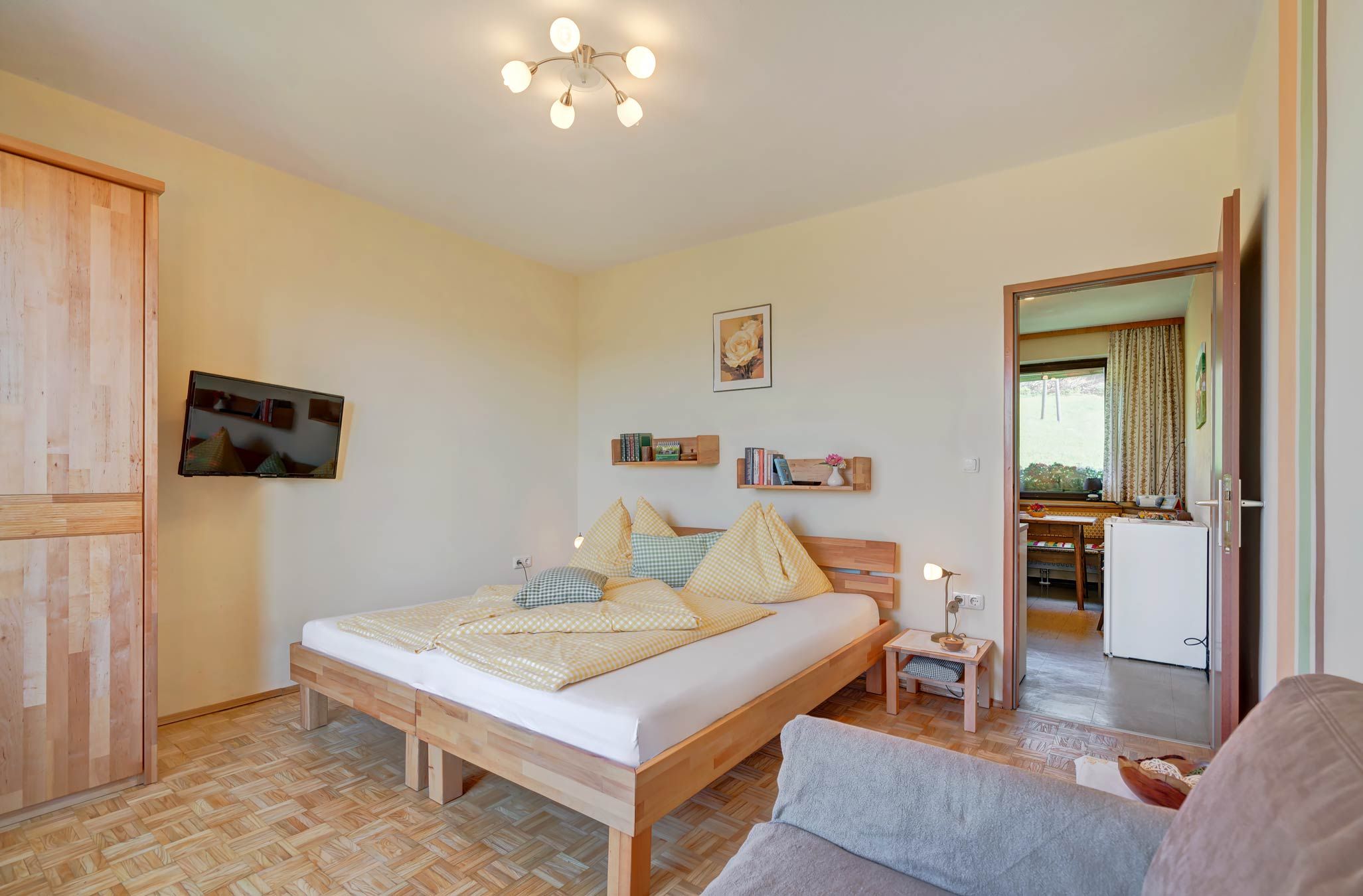 A cozy bedroom with a wooden floor, a double bed with yellow bed linen, a flat-screen TV on the wall and an open passageway to the kitchen.