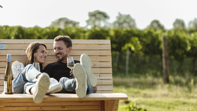 A couple relaxes on a wooden bench outside with glasses of wine in their hands.