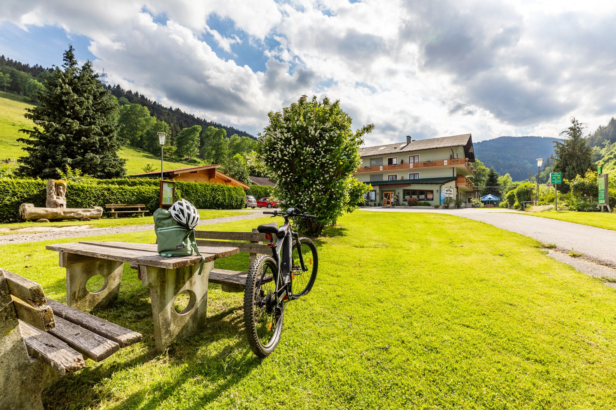 A bicycle is parked next to a picnic table on a green meadow in front of a building called Hubertushof.