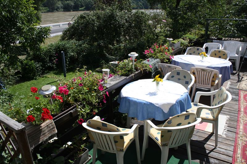 Terrace with tables and chairs, surrounded by plants and flowers, overlooking a river.