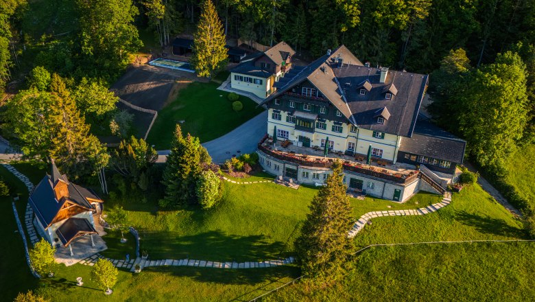Aerial view of a large building with garden and small chapel, surrounded by trees.