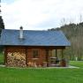 A log cabin with woodpile and barbecue in the countryside, surrounded by trees.