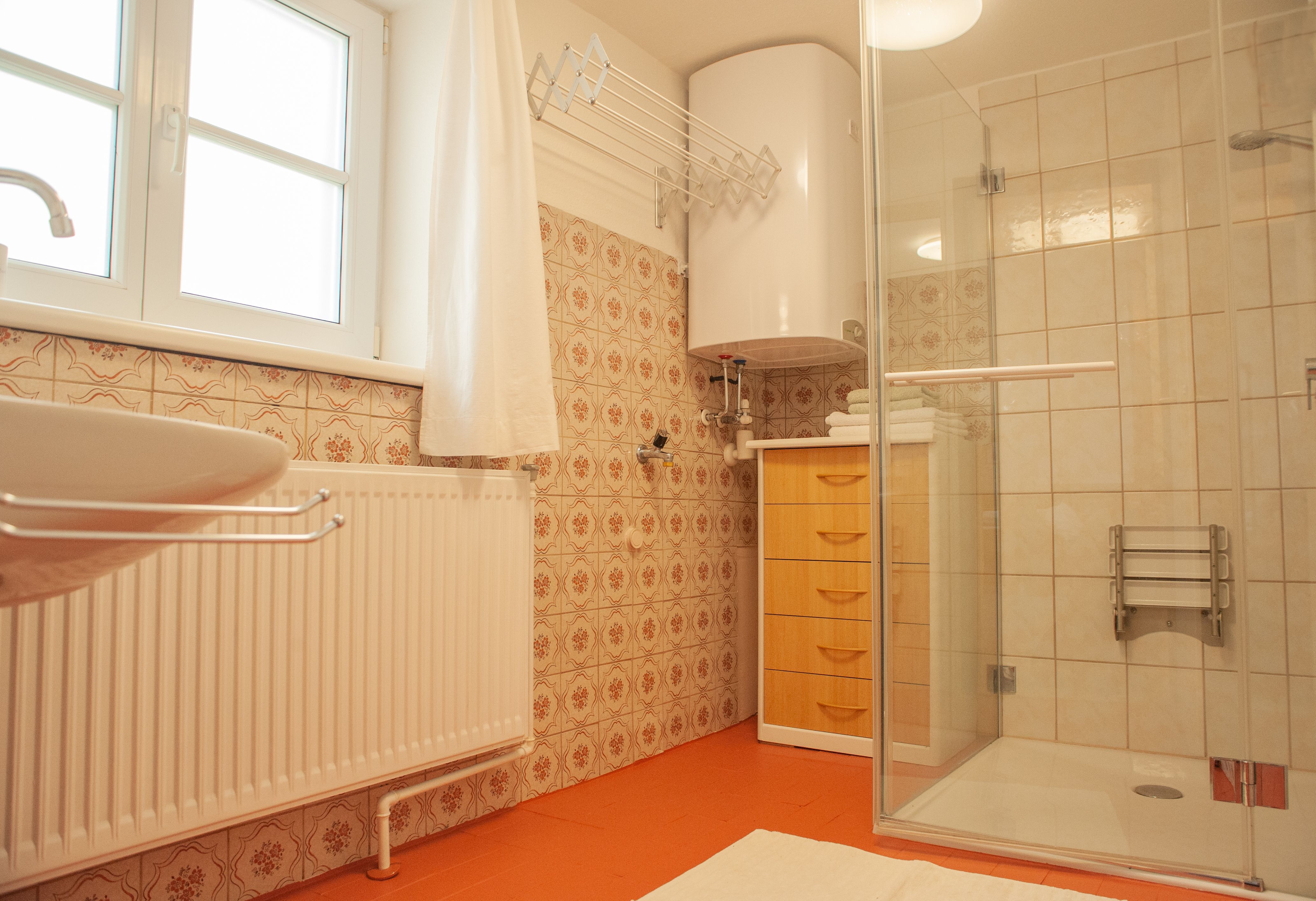 A bathroom with shower, washbasin, radiator and wooden cabinet. The walls are clad with patterned tiles.