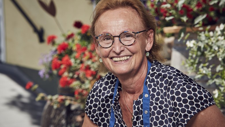 A smiling woman with glasses and a polka-dot blouse in front of flowering plants.