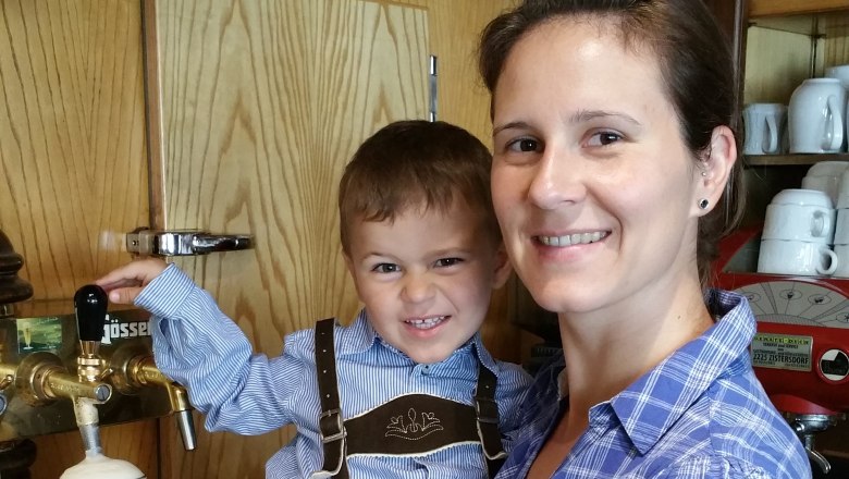 A woman holds a child in lederhosen by a beer tap. Bottles of spirits can be seen in the background.