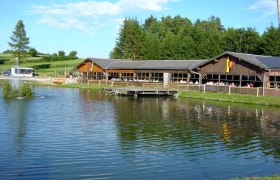 A wooden building by a pond with a fountain, surrounded by trees and meadows.