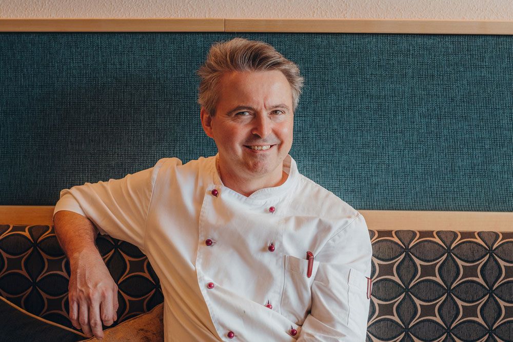 A smiling man in a white chef's jacket sits on an upholstered bench in front of a patterned wall.