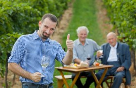 A man in a checked shirt gives a thumbs-up and holds a wine glass, while two older men sit at a table in the vineyard in the background.