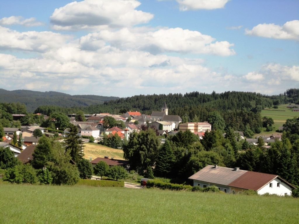 Panorama of the market town of Martinsberg with church and surrounding houses, surrounded by forests and fields under a blue sky.