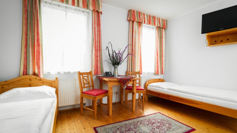 A simply furnished room with two single beds, a table with two chairs, a carpet and a TV on the wall.