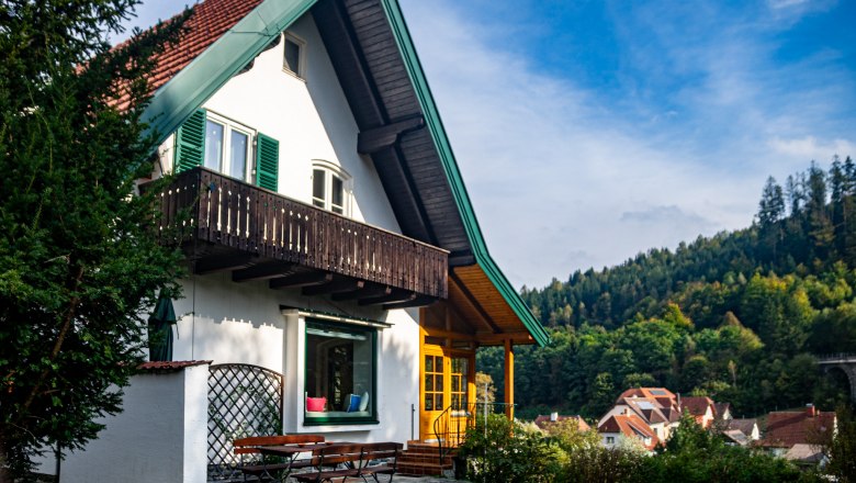 A white house with a red roof and green balcony in front of a wooded hill.